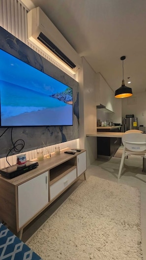 A modern living room with a flat-screen TV mounted on a wall, a wooden entertainment unit, a white sofa, a kitchen area with a dining table, and a fluffy white rug.