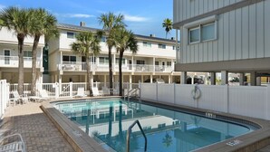 A pool area with lounge chairs and palm trees, surrounded by a multi-story building with balconies. - Gulfside Villas Unit 11... Steps to the Beach, Heated Pool Beautiful Townhome (Indian Rocks Beach)