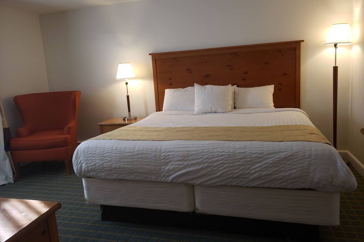 A hotel room with a large bed, a wooden headboard, two bedside lamps, a red armchair, and a small wooden table.