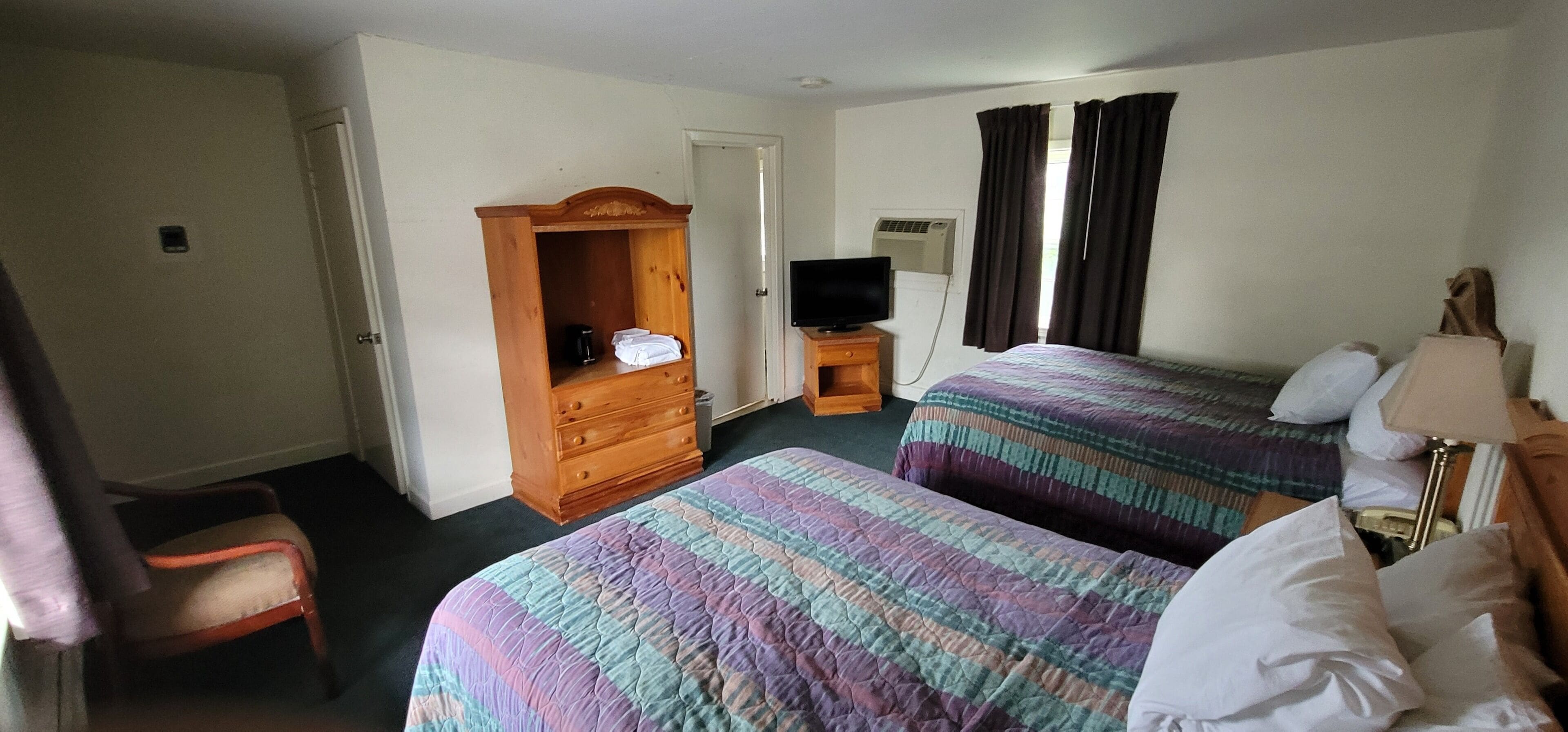 A hotel room with two beds, a wooden wardrobe, a television on a wooden stand, and a desk with a lamp.