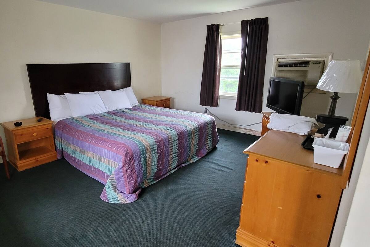 A hotel room with a bed, a chair, a nightstand, a dresser, a television, and an air conditioning unit.