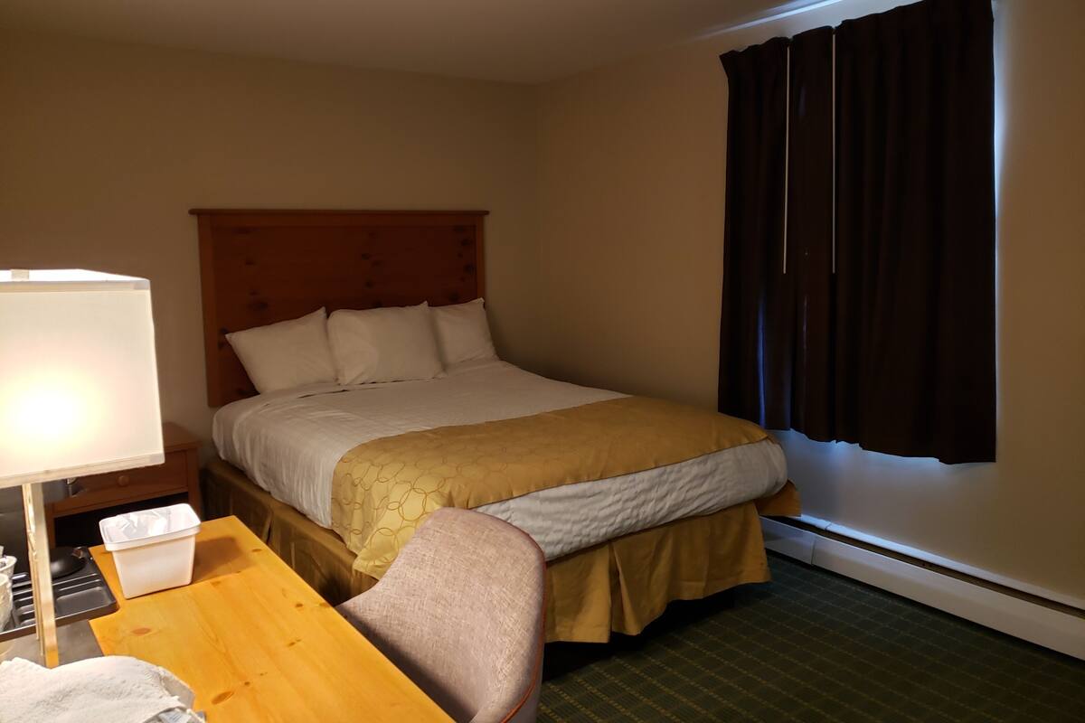 A hotel room with a bed, a bedside table with a lamp, a chair, and a window with curtains.