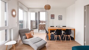 A modern living room with a grey sofa, a dining table with chairs, and a view of the outdoors through large windows.