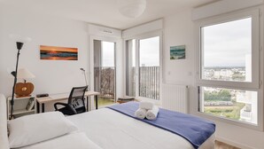 A modern hotel room with a large bed, a desk, and a view of the city through large windows.