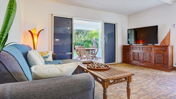 Un salon comprenant un canapé, une table basse, un téléviseur et une porte-fenêtre coulissante donnant sur une terrasse aménagée avec du mobilier d’extérieur.