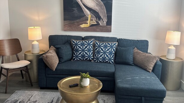 A living room with a blue sectional sofa, patterned pillows, a brass coffee table, a painting of a pelican, and a ceiling fan.