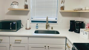 A kitchen with a microwave, coffee maker, and a sink. There are decorative items on the shelves and a window with blinds.
