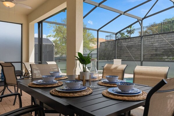 An outdoor dining area with a table set for four, surrounded by chairs, with a view of a swimming pool and a brick wall in the background.
