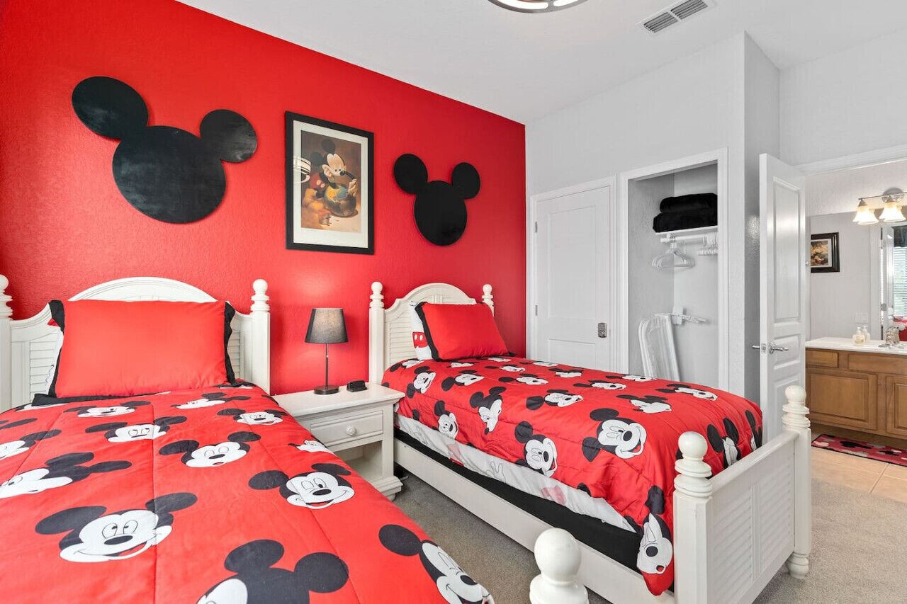 A bedroom with two beds, each with a red Mickey Mouse themed bedspread. There is a white wardrobe, a nightstand with a lamp, and a framed picture on the wall.