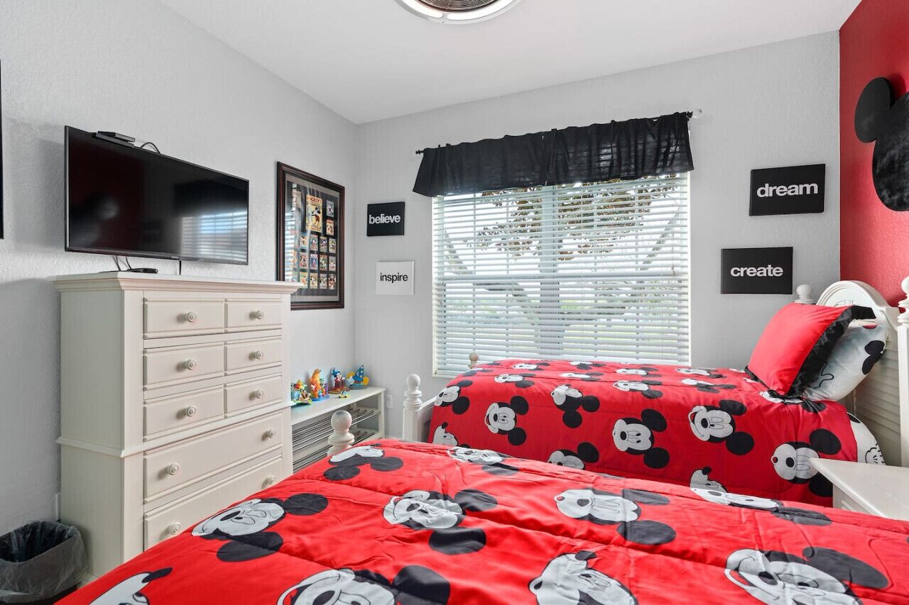 Ein Schlafzimmer mit einem Bett, das eine Mickey Mouse-Decke mit rotem Muster hat, einer weißen Kommode, einem Fernseher und einem Fenster mit Jalousien.