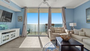 A spacious living room with a large window, a ceiling fan, a television, and a white entertainment unit. - Breathtaking Beachfront Condo, By Pier Park, Pools (Panama City Beach)