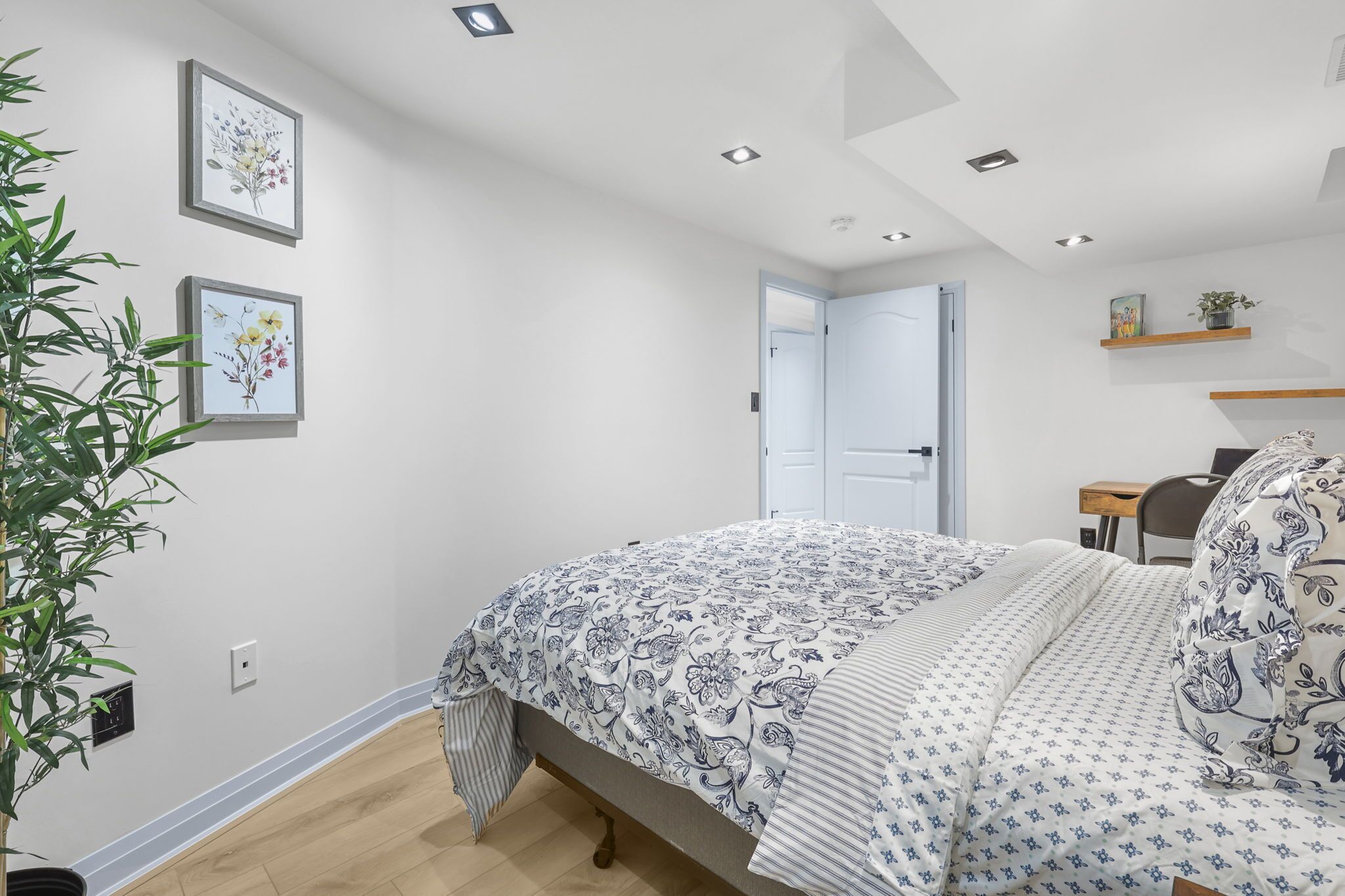 A bedroom with a bed, a bedside table, a chair, a desk, a potted plant, and framed floral artwork on the wall.