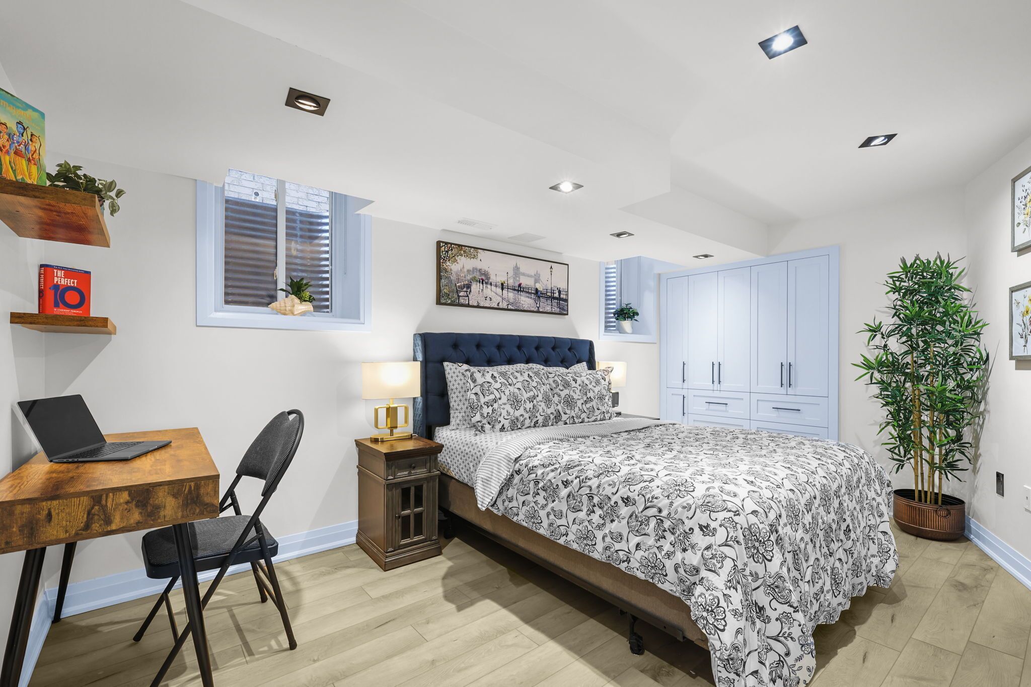 A bedroom with a bed, a desk with a laptop, a chair, a nightstand, a window, and a potted plant.