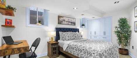 A bedroom with a bed, a desk with a laptop, a chair, a nightstand, a window, and a potted plant.