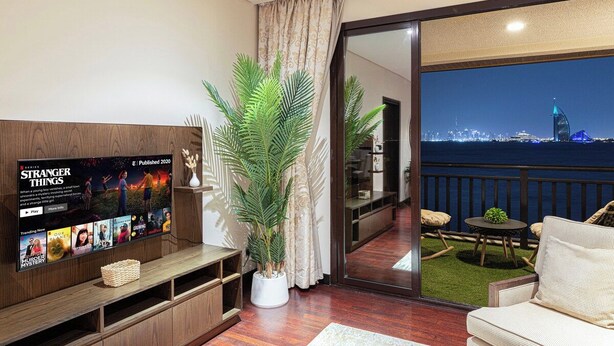 Un salon moderne avec un téléviseur à écran plat, une plante en pot et un balcon offrant une vue sur les toits illuminés de la ville la nuit.