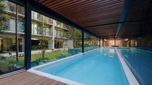 Een modern binnenzwembad met een houten terras, glazen wanden en uitzicht op een woongebouw.