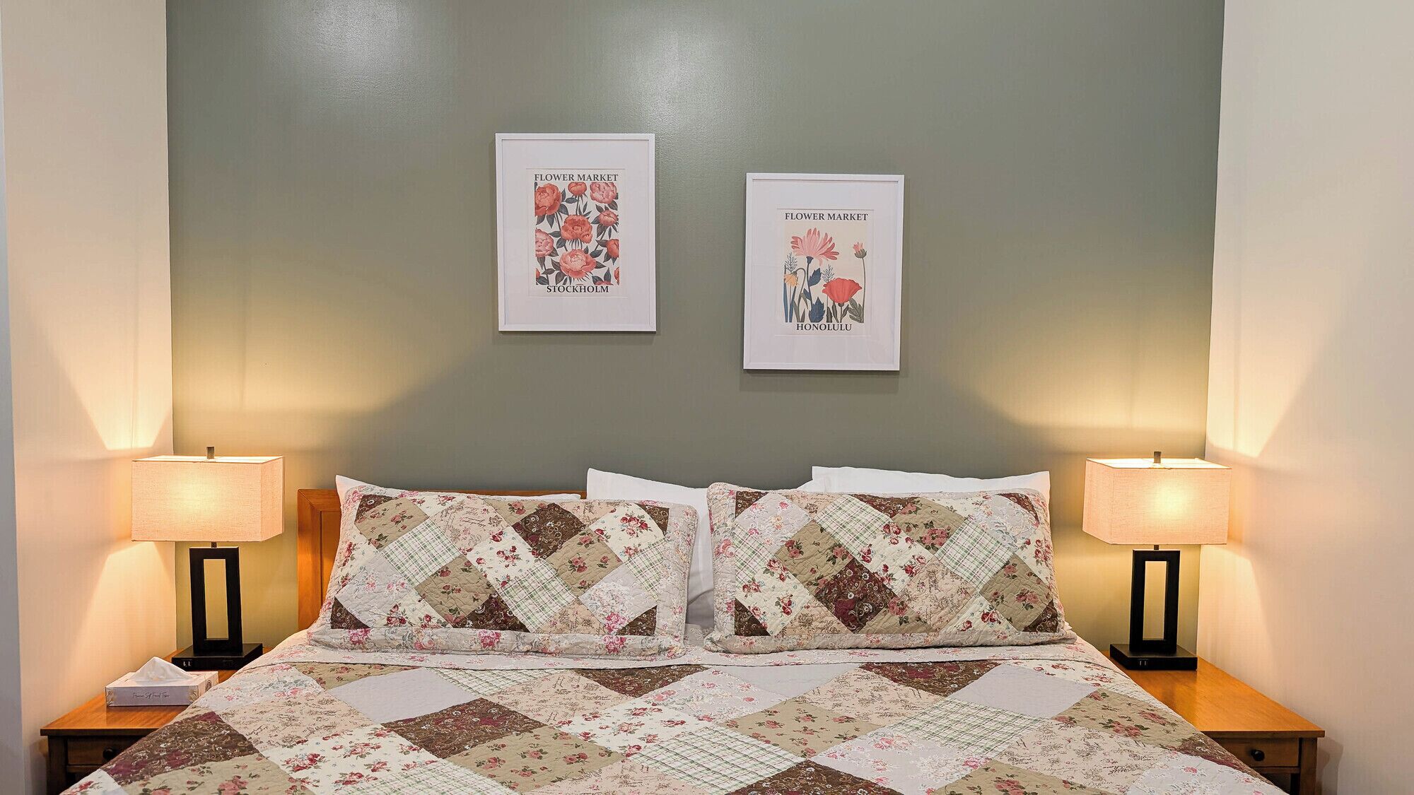 A bedroom with a floral quilt, two framed floral artwork on the wall, and bedside lamps.