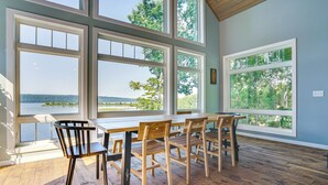 Ein Essbereich mit großem Tisch und Stühlen, großen Fenstern mit Blick auf einen See und Bäume, sowie Holzboden.