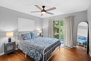 Une chambre à coucher avec un lit, deux tables de chevet avec des lampes, un ventilateur de plafond, une grande fenêtre avec des rideaux et une vue sur l’extérieur.