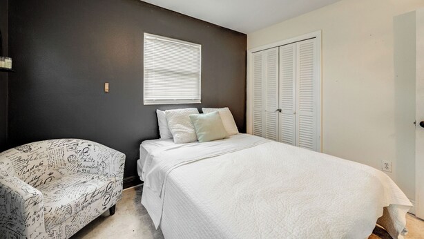 A bedroom with a bed, a nightstand, a chair with a patterned cover, a window with blinds, and a door. - Orange Grove (Melbourne)