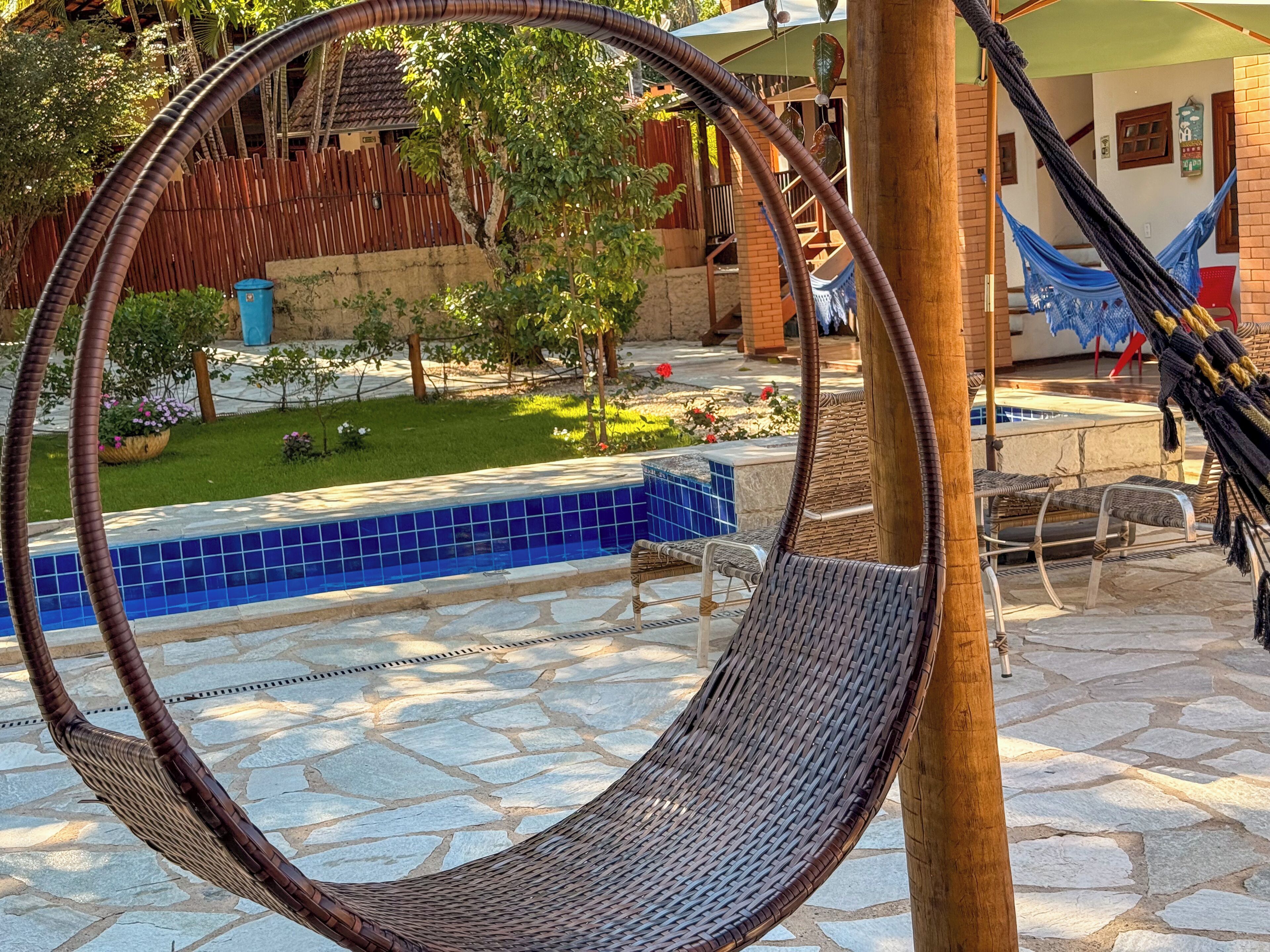 Un hamac est suspendu entre deux poteaux en bois, dans une cour où se trouve une piscine carrelée, du mobilier d’extérieur et, au fond, une clôture en bois.