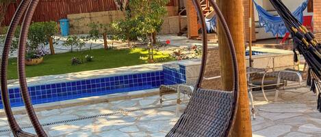 Un hamac est suspendu entre deux poteaux en bois, dans une cour où se trouve une piscine carrelée, du mobilier d’extérieur et, au fond, une clôture en bois.