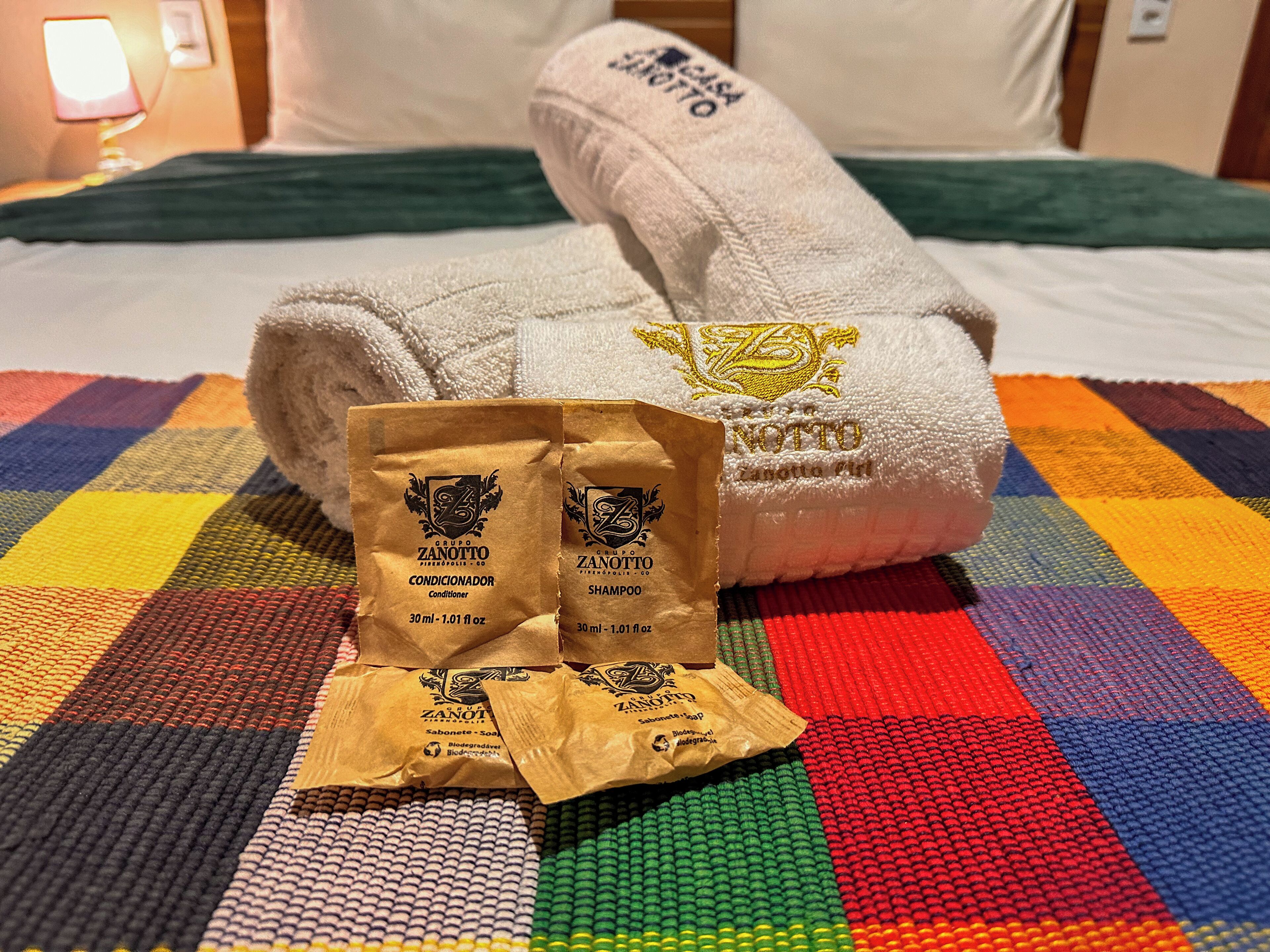A bed with a colorful checkered bedspread, two towels, and two bags of toiletries.