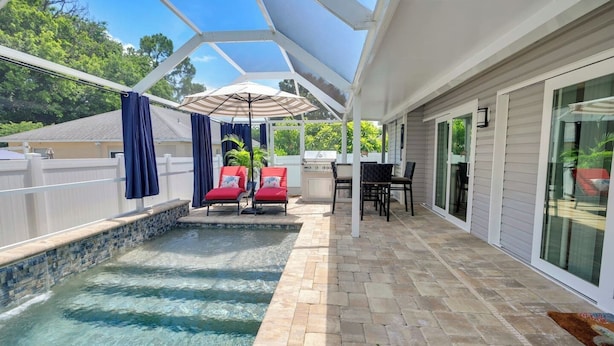 Un área de piscina exterior techada con sillas de descanso y una mesa, rodeada por una cerca blanca y un muro de piedra.