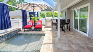 Un patio techado al aire libre con piscina, sillas reclinables rojas, una parrilla y una mesa con sillas.