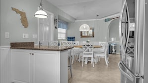 Una cocina con muebles blancos, encimera de granito, una zona de comedor con sillas blancas y un refrigerador de acero inoxidable.