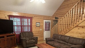 A cozy living room with a wooden staircase, leather furniture, and a flat-screen TV. - Bear-A-Dise- A beautiful mountain cabin-sleeps 12, hot tub, game room & firepit. (Blairsville)