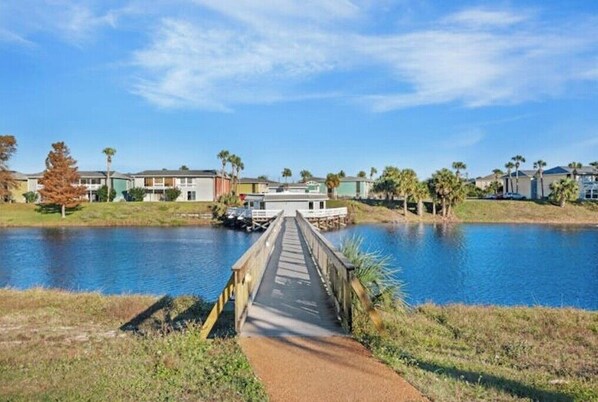 A wooden bridge over a calm body of water, leading to a row of houses with a clear blue sky above.