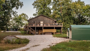 A rustic wooden cabin with a porch and a garage, surrounded by trees and a gravel path. - 4BR, 3.5 BA House, great sunset view, close to town. (Nashville)