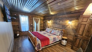 A cozy bedroom with a wooden bed, a bedside table, and a chair. There is a window with curtains and a door with a towel rack. - 4BR, 3.5 BA House, great sunset view, close to town. (Nashville)