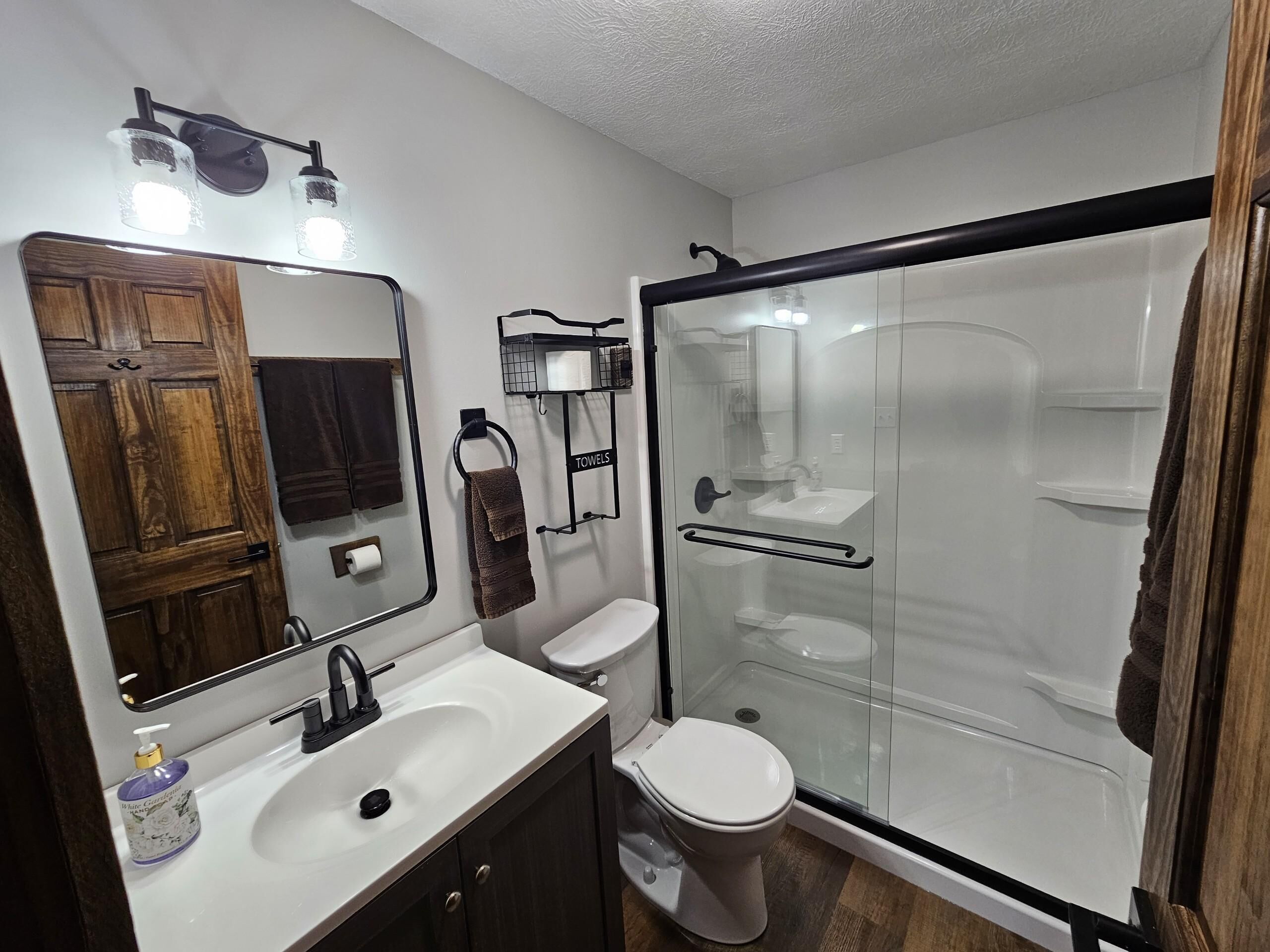A modern bathroom with a wooden vanity, a toilet, a glass-enclosed shower, and a mirror with lights above the sink.