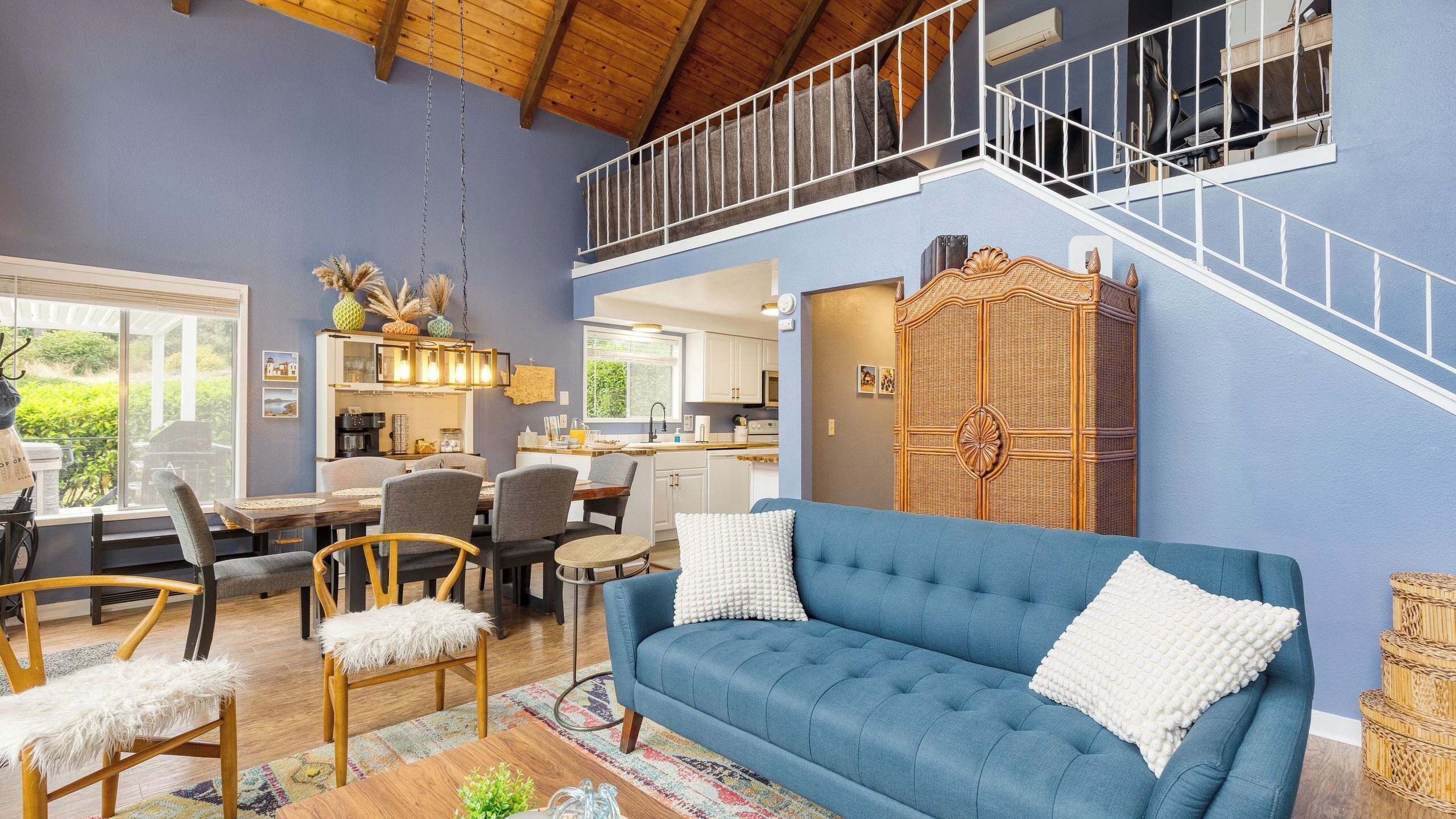 Un vaste espace de vie comprenant un canapé bleu, une table à manger et des chaises. On aperçoit une cuisine au fond, et un escalier mène à l’étage supérieur.
