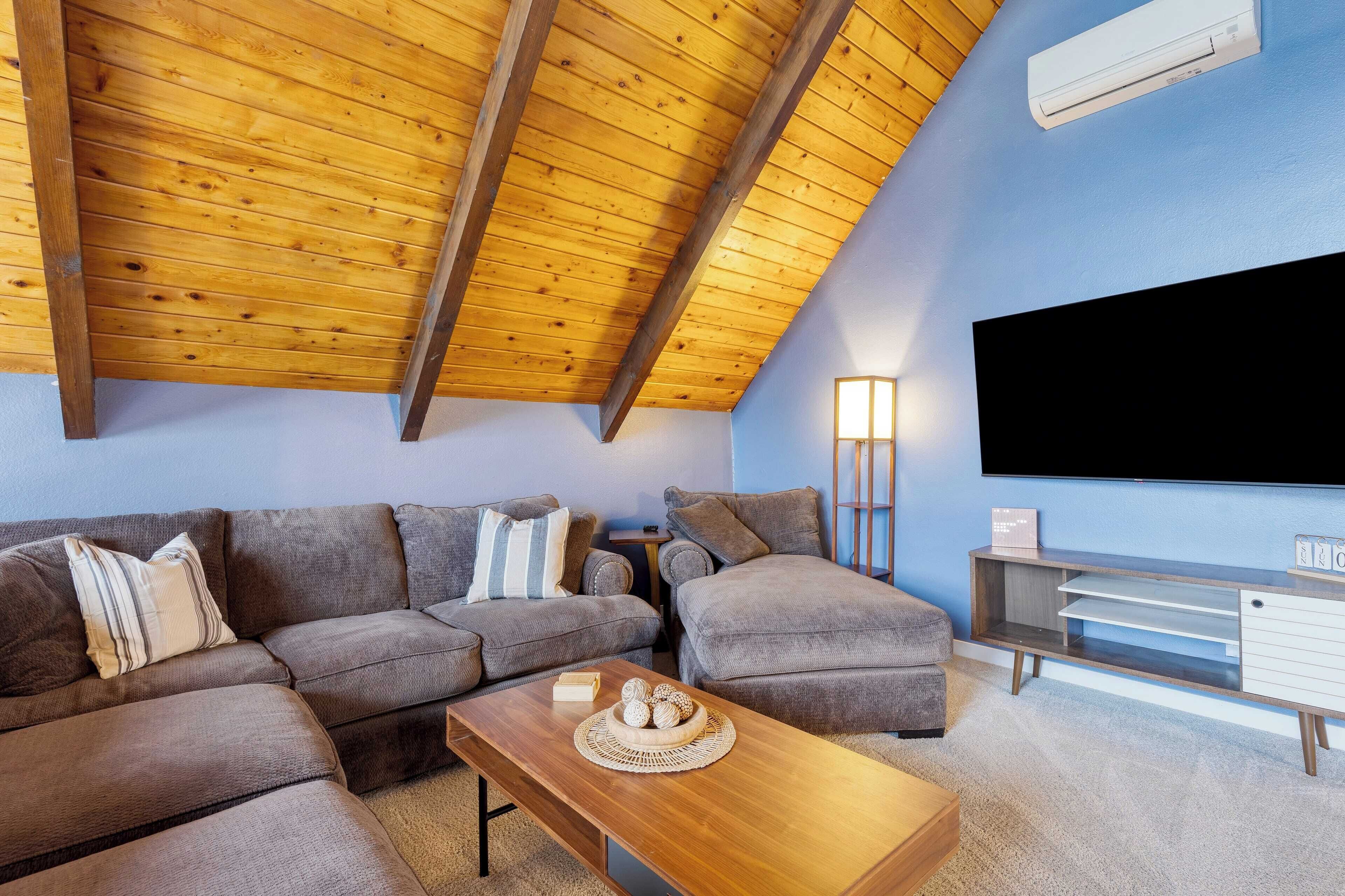 A living room with a wooden ceiling, grey sofas, a wooden coffee table, and a flat-screen TV mounted on the wall.