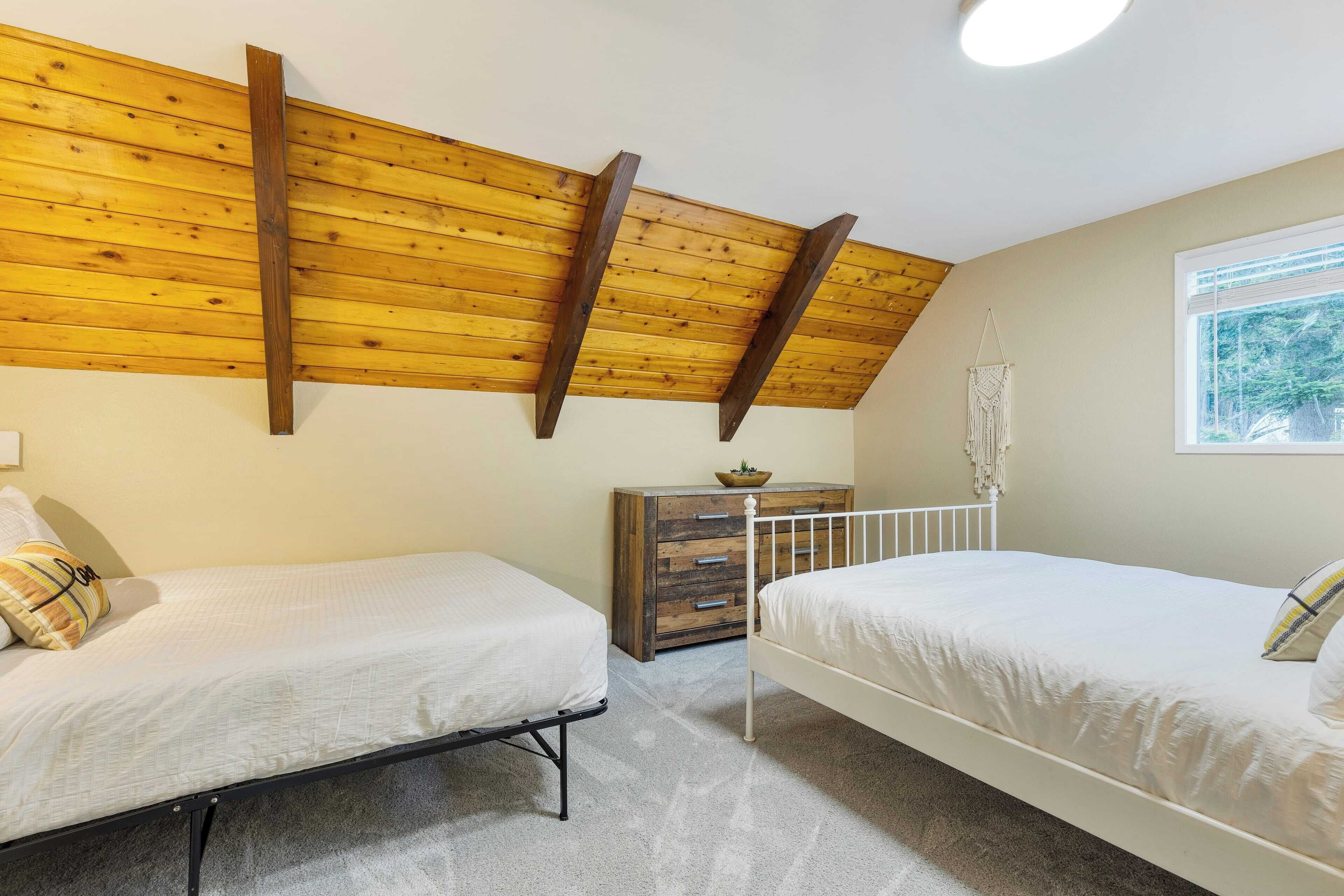 A bedroom with two single beds, a wooden ceiling, a dresser, and a window with a view of greenery.