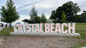 Letrero que dice 'CRYSTAL BEACH' con un fondo de árboles y un cielo nublado.