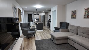 Una sala moderna con un sofá gris, dos sillones y una mesa de centro. Se ve una cocina al fondo, con armarios blancos y electrodomésticos de acero inoxidable.