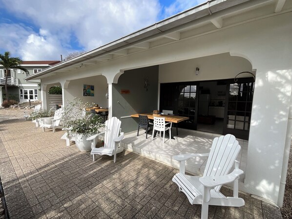 A covered patio area with white Adirondack chairs and a wooden table, featuring a view of the house and a door leading to the kitchen. - Apartment on gated resort, minutes from beautiful beaches (Sint Michiel)