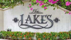 Una pared con el texto "The Lakes" en letras decorativas, rodeada de arbustos verdes y flores rosadas.