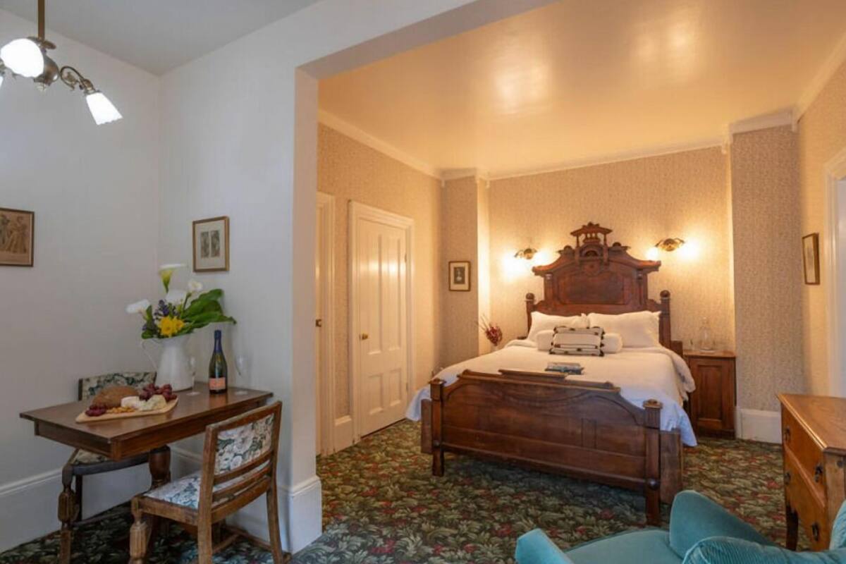 Un dormitorio con una cama de madera, un escritorio con silla, un florero con flores y piso alfombrado.