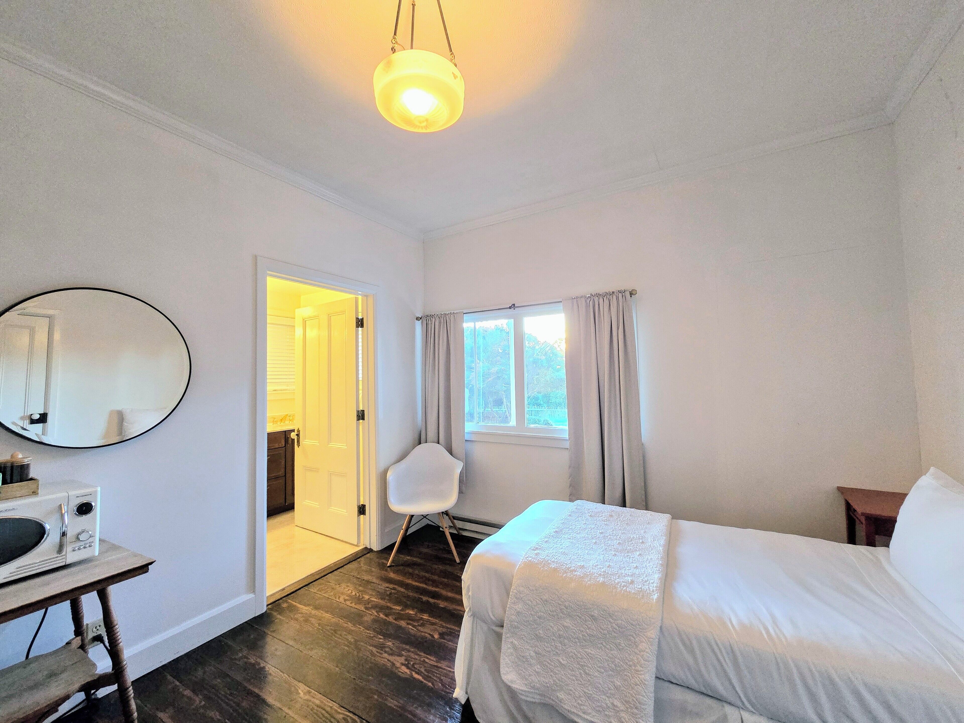 Habitación de hotel con una cama, un escritorio con microondas, una silla, una ventana con cortinas y una puerta que da a otra habitación.