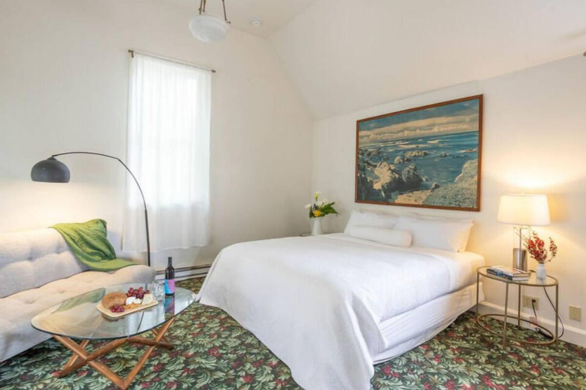 Una habitación de hotel con cama, sofá, una mesa de vidrio con frutas, lámpara y un cuadro en la pared.