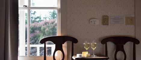 A room with a window, two wooden chairs, a small table with two glasses of wine and a piece of cheese.
