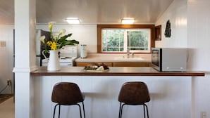 A modern kitchen with white cabinetry, a wooden countertop, two bar stools, a window with a view of greenery, and a refrigerator.