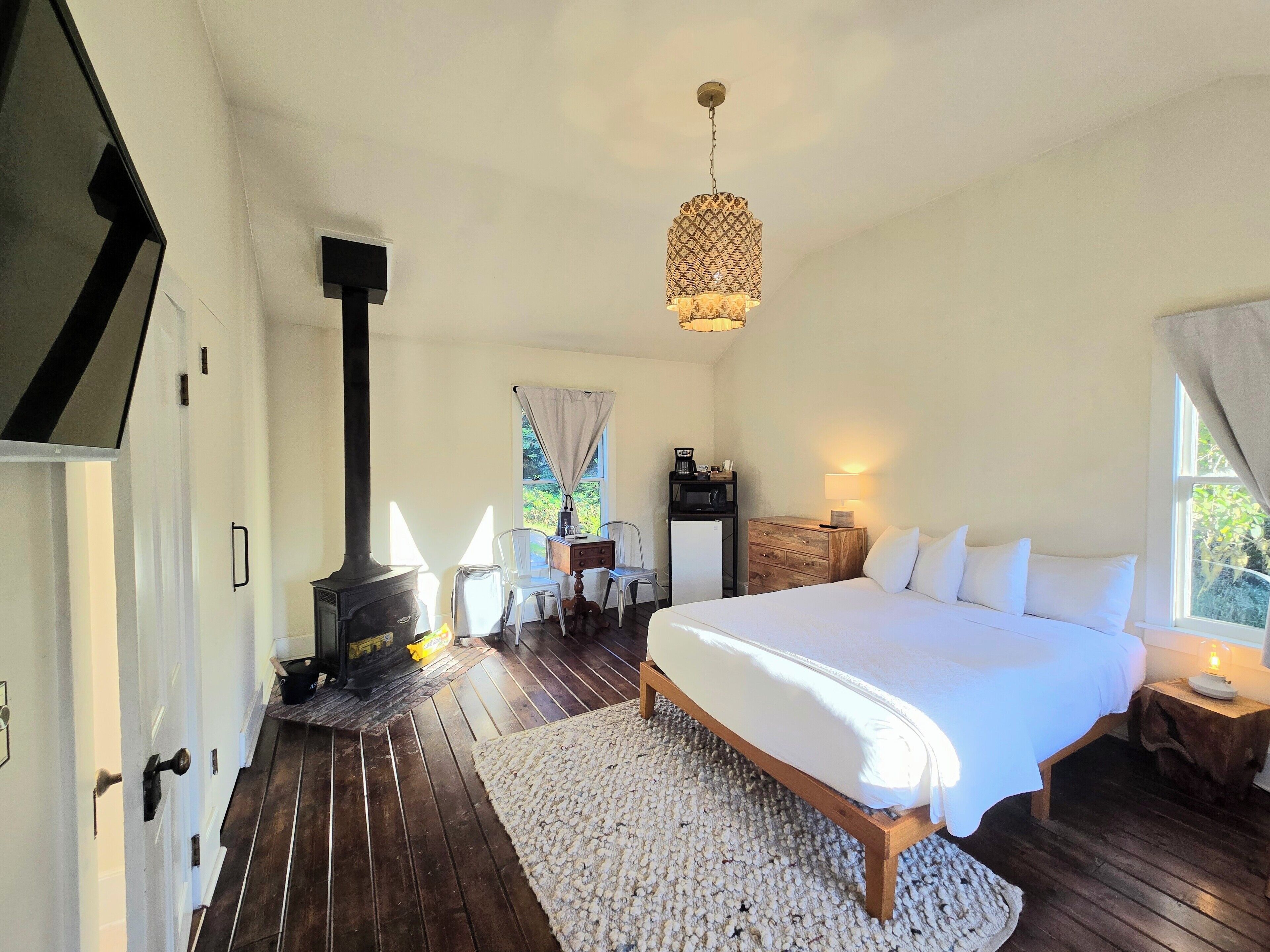 A bedroom with a large bed, a wooden floor, a fireplace, a desk with a chair, a lamp, and a window with curtains.