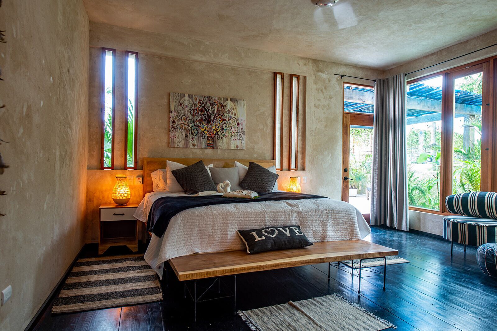A bedroom with a bed, bedside lamps, a bench, a large window, and a view of greenery.