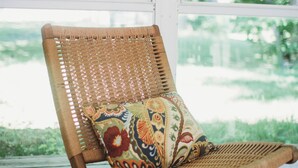 Una silla de mimbre con un cojín colorido, ubicada en una terraza de madera con vista a la vegetación a través de un ventanal grande.